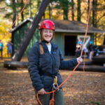 Student Ahna Webster stands at the base of the Alpine Tower challenge course holding ropes and smiling.