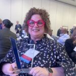 Dr. Lucy Lawrence stands at the PACE Conference holding the Engaged Faculty Award. She is wearing bright red glasses and a polka dot shirt.