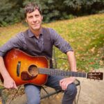 Professor John Cloyd Miller sits outside holding his guitar posed for a photo.