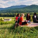 Students sit on dogwood pasture speaking with one another with the backdrop of mountains and the white barn.