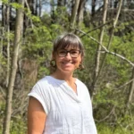 Professor Kristen Keane smiles for a headshot in front of a wooded backdrop.
