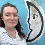 Visiting Assistant Professor of Creative Nonfiction, Bailey Pittenger, PhD, stands and smiles in front of a mural of a moon.